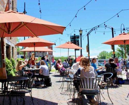 Outdoor café scene with patrons sitting under orange umbrellas, enjoying food and drinks on a sunny day.