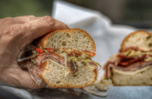 A hand holding a sandwich filled with layers of meats, lettuce, and tomatoes, with another sandwich in the background.
