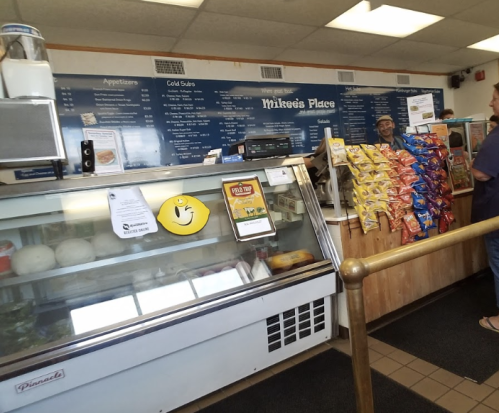 A deli counter with a menu board, snacks on display, and a smiling employee serving customers.