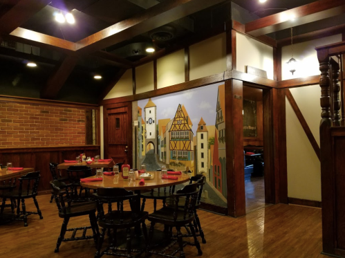 Cozy restaurant interior with wooden beams, brick walls, and a mural of a quaint village scene. Tables set for dining.