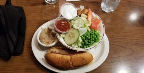 A plate with a hot dog, a side of mashed potatoes, and a salad with lettuce, tomatoes, cucumbers, peas, and dressing.