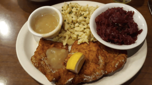A plate featuring breaded fried fish with lemon, served with spaetzle, red cabbage, and a side of gravy.