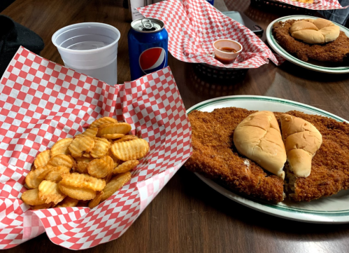 A plate with a large fried chicken cutlet, two buns, and a side of wavy potato chips, with drinks on the table.
