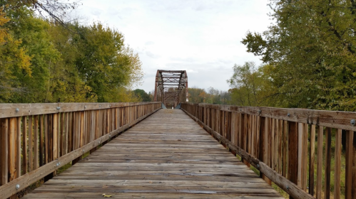 A wooden bridge stretches into the distance, surrounded by trees with autumn foliage under a cloudy sky.