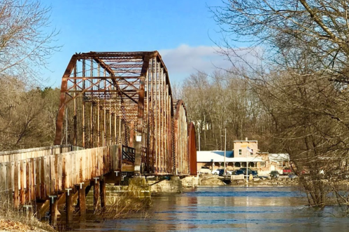 Rusty bridge over a river, with trees in the background and a building visible on the shore. Clear blue sky above.