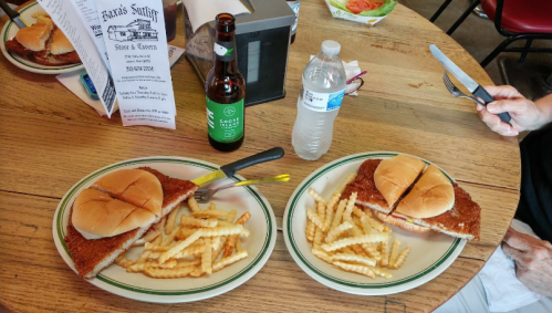Two plates with breaded sandwiches, fries, a bottle of beer, and a water bottle on a wooden table.