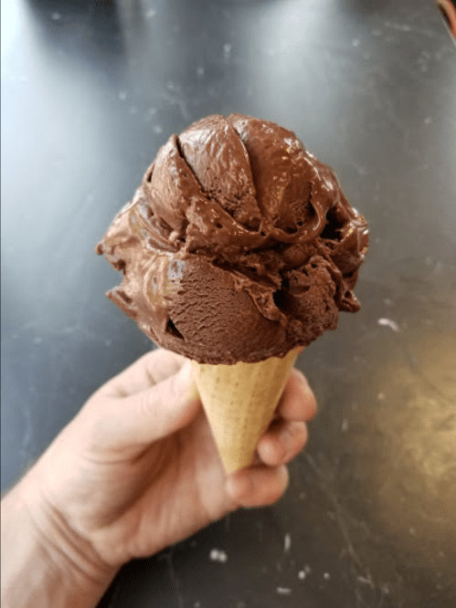 A hand holding a large chocolate ice cream cone with multiple scoops on a dark surface.