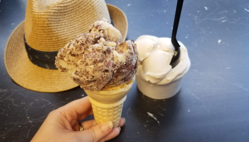 A hand holding a cone with chocolate chip ice cream, next to a cup of vanilla ice cream and a straw hat in the background.