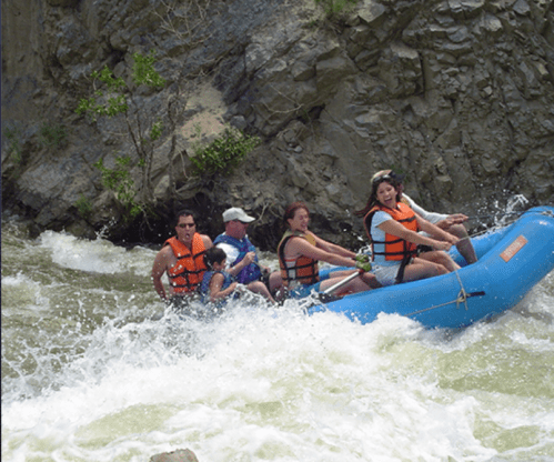 A group of people in life jackets navigating a river rapids in a blue raft, splashing through the water.