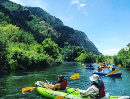 A group of people kayaking on a river surrounded by lush green mountains under a clear blue sky.