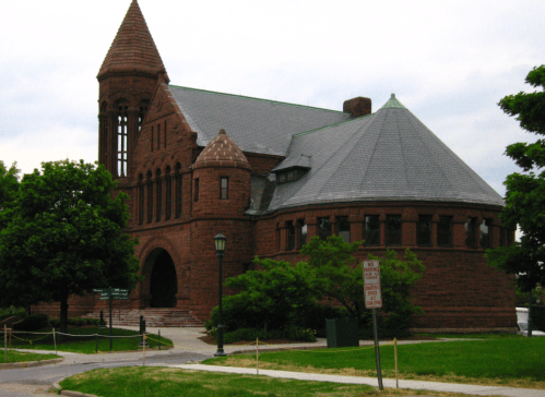 Historic red stone building with a pointed tower, surrounded by trees and a grassy area.