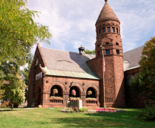 Historic red stone building with a turret, surrounded by greenery and flowers, under a clear blue sky.