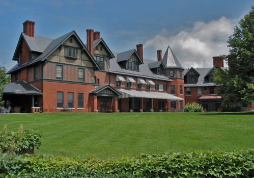A large, historic mansion with a mix of brick and stone, surrounded by green lawns and trees under a blue sky.