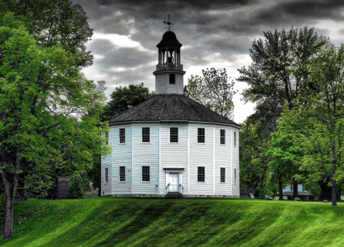 A round white building with a bell tower, surrounded by lush green trees under a cloudy sky.