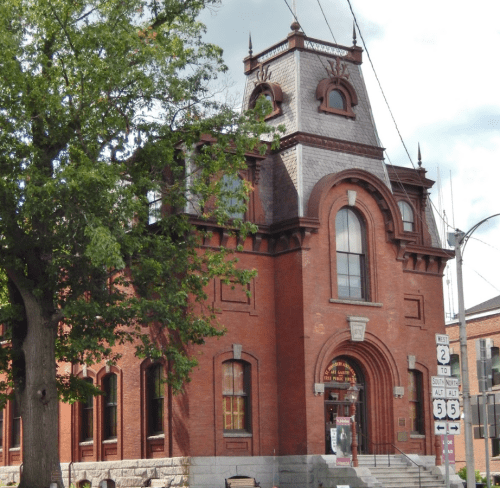 Historic red brick building with a tower, ornate windows, and a large entrance, surrounded by trees and street signs.