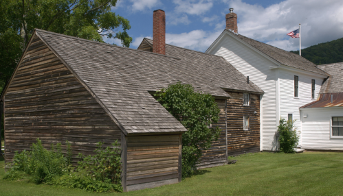 A rustic wooden building next to a white house, with green grass and trees under a blue sky with clouds.