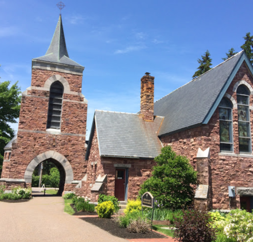 A stone church with a tall spire and a smaller adjacent building, surrounded by greenery under a clear blue sky.