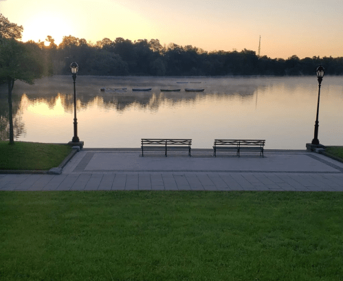 A serene lakeside view at sunrise, featuring benches, mist over the water, and boats in the distance.