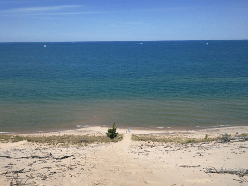 A sandy dune overlooking a calm blue lake with a few sailboats in the distance and people walking along the shore.