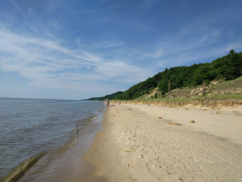 A sandy beach stretches along a calm lake, with green hills in the background and a clear blue sky above.