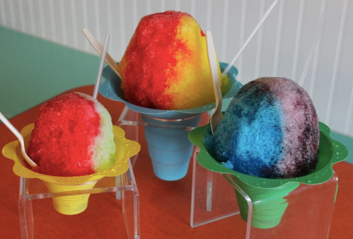 Three colorful snow cones in flower-shaped cups: red, yellow, and blue, with plastic spoons, on a bright table.