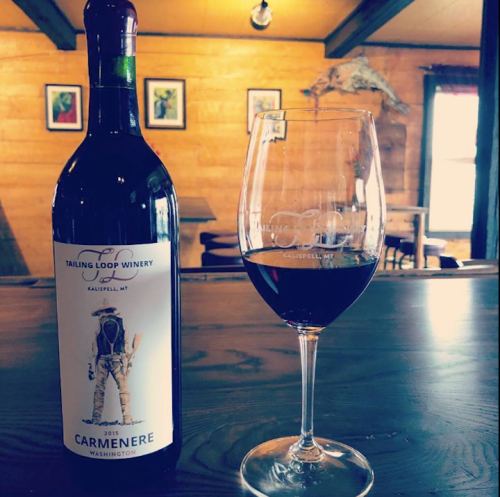 A bottle of Tailing Loop Winery 2015 Carménère next to a glass of red wine on a wooden table in a cozy setting.