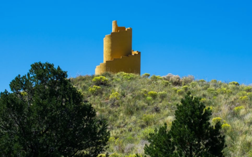A yellow tower stands atop a grassy hill, surrounded by green shrubs and trees under a clear blue sky.