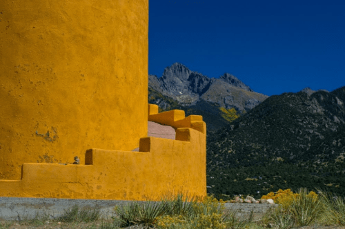A vibrant yellow building with a textured wall, set against a backdrop of rugged mountains and a clear blue sky.