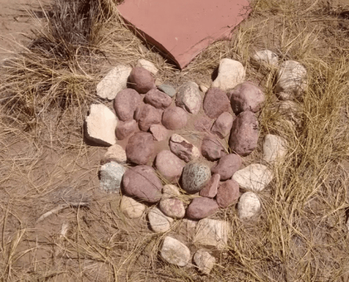 A heart shape made of various stones, surrounded by dry grass and a clay heart in the background.