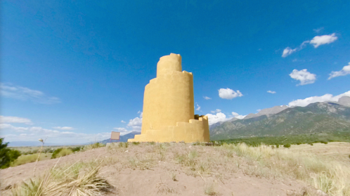 A tall, yellow adobe structure stands on a hillside with mountains and a blue sky in the background.