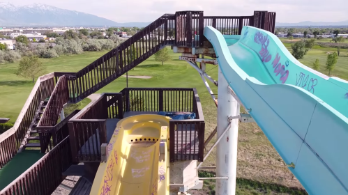 Aerial view of a water slide structure with a yellow and turquoise slide, surrounded by green grass and trees.