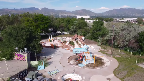 An aerial view of an abandoned water park with overgrown vegetation and damaged slides against a mountain backdrop.