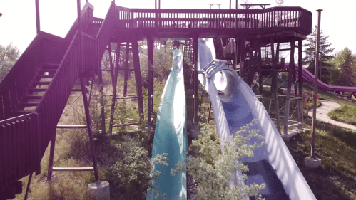 Two water slides in an amusement park, one blue and one green, with a wooden structure and stairs in the background.