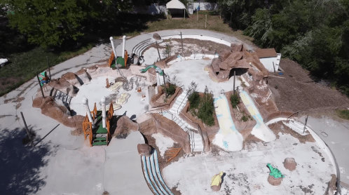 Aerial view of an abandoned playground with slides, rock formations, and overgrown greenery.