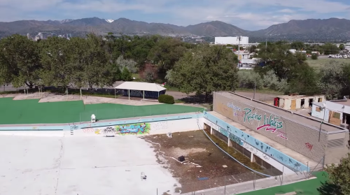 Aerial view of an abandoned pool surrounded by graffiti, with mountains and trees in the background.