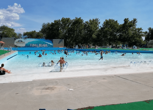 A crowded water park with people swimming and playing in a large pool on a sunny day.