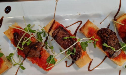 A plate of sliced bread topped with tomato sauce, beef, and microgreens, drizzled with balsamic glaze.