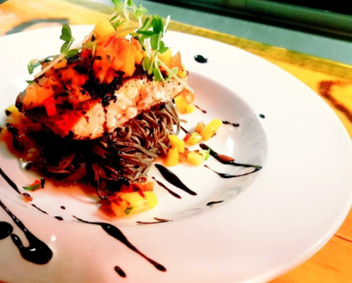 A beautifully plated dish featuring grilled fish atop soba noodles, garnished with mango and drizzled with sauce.