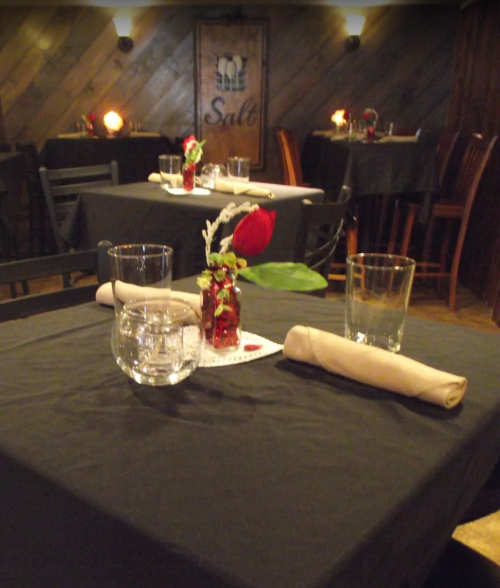 A cozy restaurant interior with black tablecloths, glassware, and a small vase with a rose on each table.