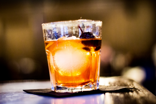 A glass of amber-colored cocktail with ice and a cherry, set on a dark surface with a blurred background.