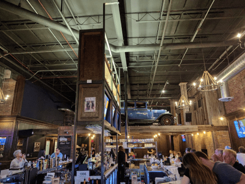 A lively bar interior with a vintage car on display above, wooden accents, and patrons enjoying their time.