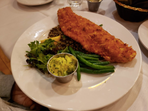 A plate with a crispy fish fillet, green beans, mixed greens, and a side of sauce, served on a white tablecloth.