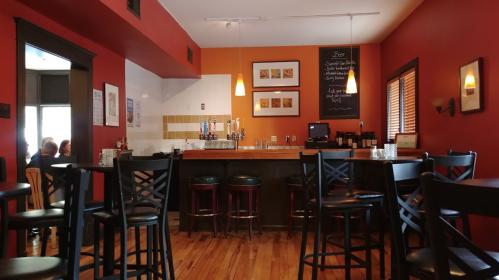 Cozy bar interior with wooden floors, a counter, stools, and colorful walls. A chalkboard menu is visible.