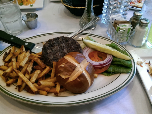 A plate with a burger, pretzel bun, lettuce, tomato, onion, pickles, and a side of fries.