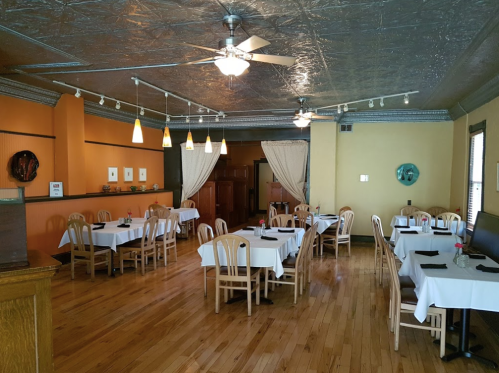 Cozy restaurant interior with wooden floors, white tablecloths, and warm orange walls, featuring ceiling fans and soft lighting.