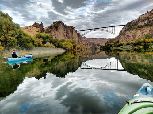 A person kayaking on a calm river, with a bridge and rocky cliffs reflected in the water under a cloudy sky.