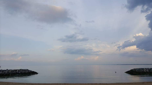 Serene beach scene with calm water, distant rocky outcrops, and a cloudy sky reflecting soft pastel colors.