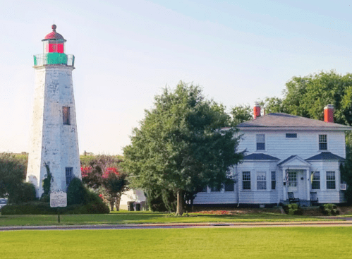 A white lighthouse with a green and red top next to a white house, surrounded by trees and a clear blue sky.