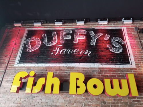 Sign for Duffy's Tavern with a bright yellow "Fish Bowl" sign below, set against a brick wall.
