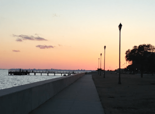 A serene waterfront at sunset, with a pier and lampposts lining the path along the shore.